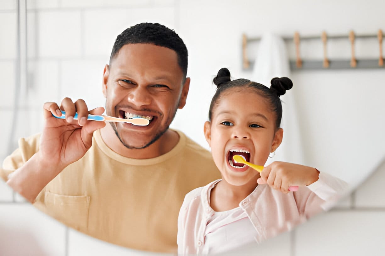 Father and daughter smiling and brushing their teeth together in front of a bathroom mirror, demonstrating the importance of family oral hygiene and healthy dental habits