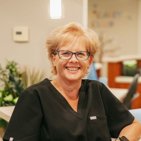 Headshot of Beth Jones, Denture Technician with short blonde hair and glasses poses at the front desk of a modern dental office