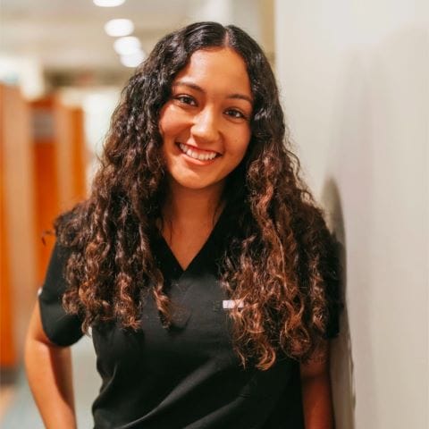 Headshot of Alma Gonzalez, Dental Hygienist with long curly hair stands in a bright dental office hallway
