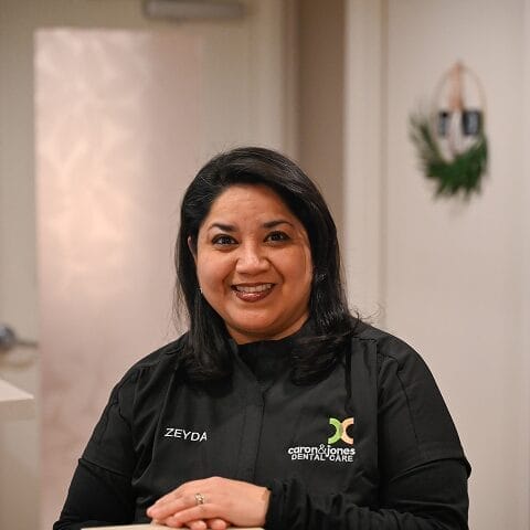 Headshot of Breana Brown, Insurance Coordinator in black scrubs smiles at the camera while sitting at a dental office front desk, conveying professionalism and approachability