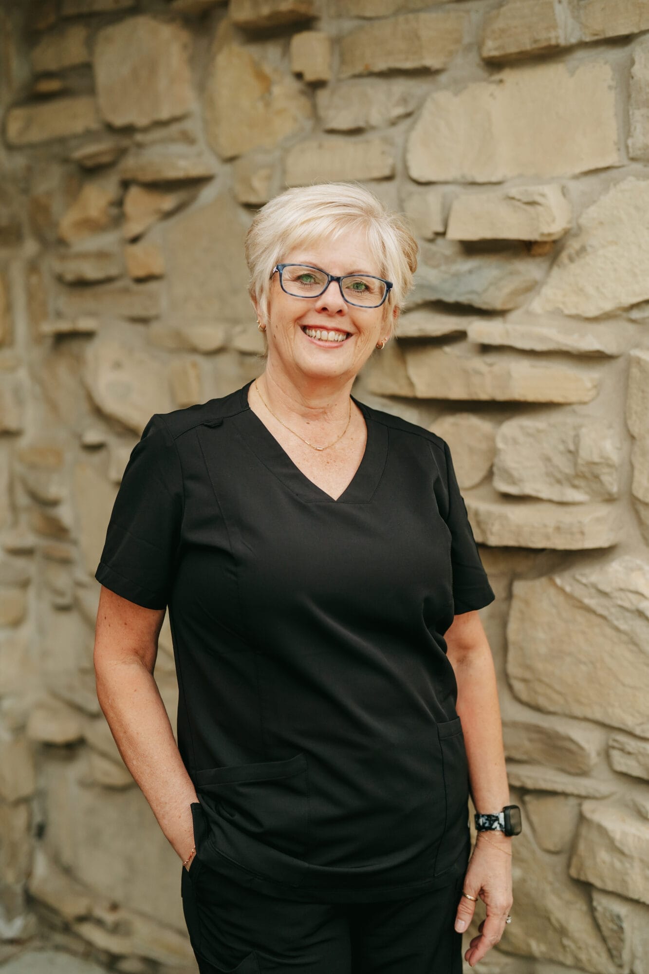 Headshot of Beth Jones, Denture Technician with short blonde hair and glasses poses at the front desk of a modern dental office